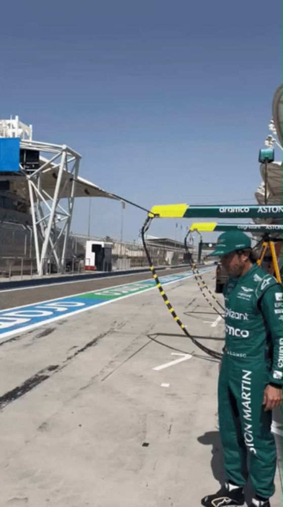 a man wearing a green aston martin jumpsuit stands in a pit lane