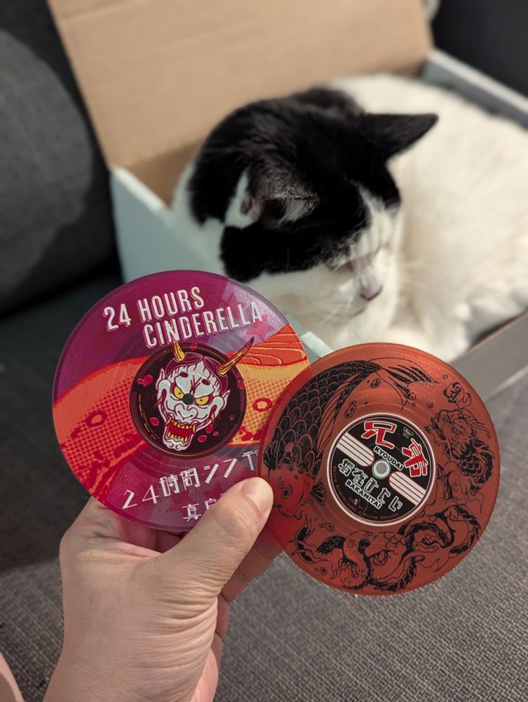 Two plastic coasters shaped like music compact disks, decorated to celebrate songs from the Yakuza franchise: 24 Hours Cinderella for Majima Goro and Bakamitai for Kazama Kiryu.