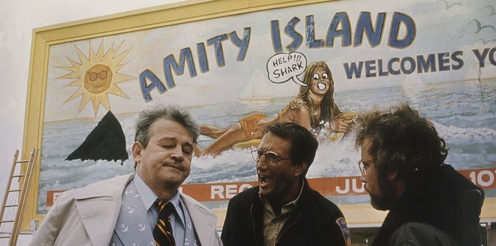 A wooden sign saying Amity Island, welcomes you, with a picture of a beach scene. Three men stand in front.
