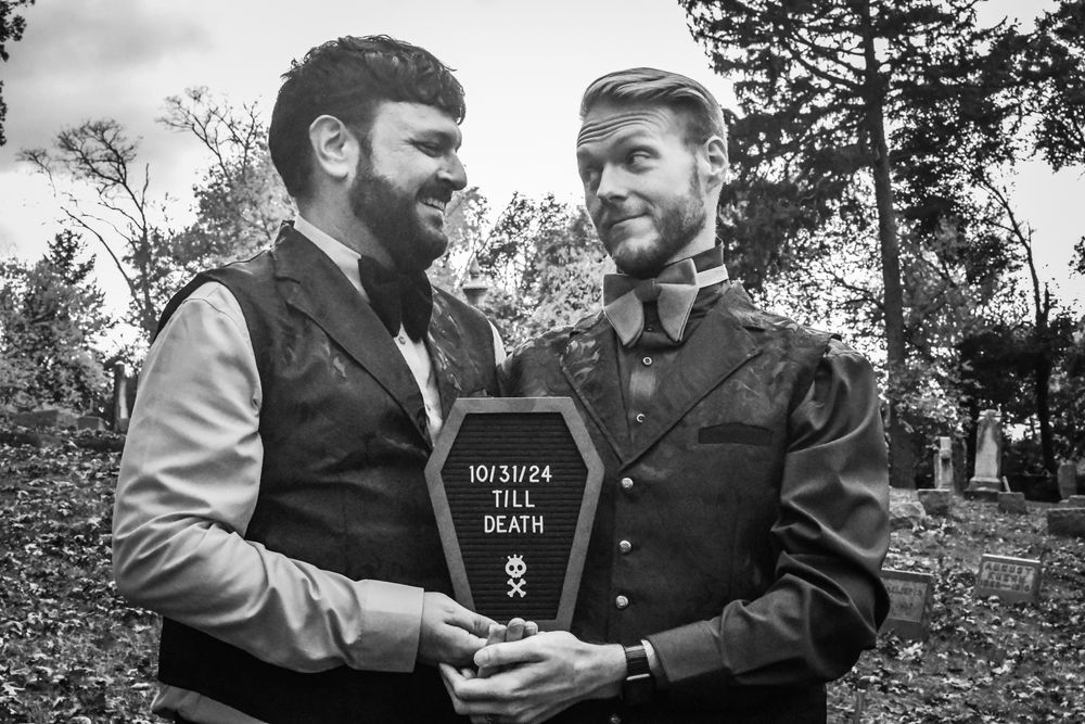 Black and white photo of Sean and Tim in a cemetery holding a coffin shaped sign that says “10/31/24 - till death”