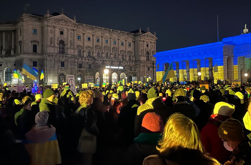 Vienna's Heldenplatz at night. A crowd of thousands stands in the plaza waving flags and signs, surrounded by neoclassical buildings. The blue and yellow stripes of the Ukrainian flag are projected on to the historic Burgtor (monumental gate). 
