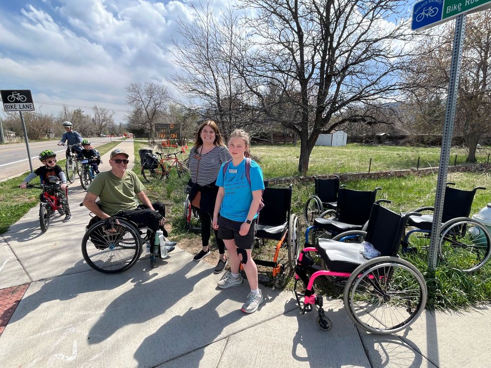 Three people on a street corner surrounded by empty wheelchairs. One of the people is seated in a wheelchair. An adult and two kids approach on bikes on the sidewalk.