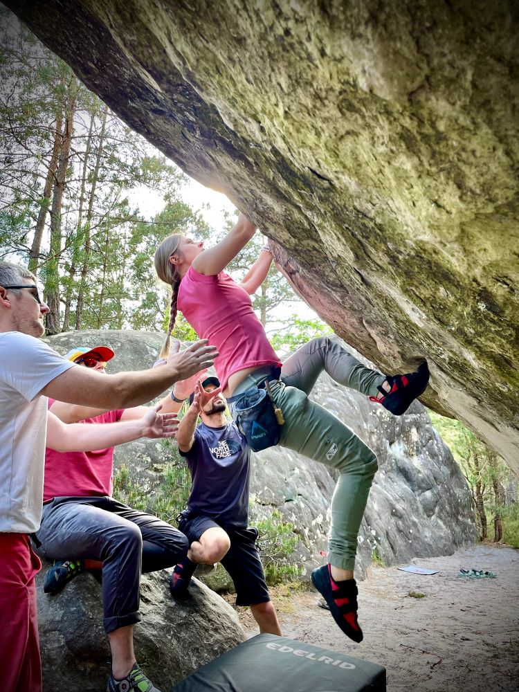 Emma climbing an overhanging boulder in the forests of Fontainebleau with three people spotting.