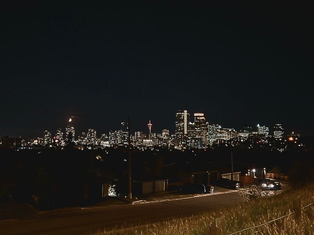 A photo of the downtown Calgary skyline at night, with a crescent moon in the sky and some garages and grass in the foreground 