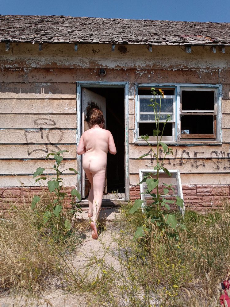 Hayley Seas is pictured in this portrait walking into an abandoned house. The sun is bright illuminating the stark colors of overgrown green foliage around the house. A wild flower taller than her is bright orange. Her body reflects the sun as if it were meant to be used that way. Hayley Seas body expresses some unique folds and curves that only a photographer can capture with a plus size body. 
