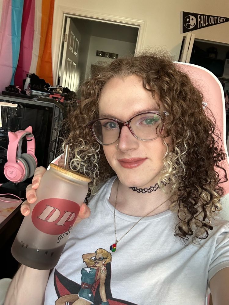 selfie of kara smiling slightly and holding up a frosted glass tumbler with "DP girlie" on the front like the Dr Pepper logo. her makeup is expertly applied with glossy lips and stunning eyeliner and her curly hair is catching the light just divinely. she's wearing large round glasses a tattoo choker and a gold necklace with a color by addition venn diagram pendant