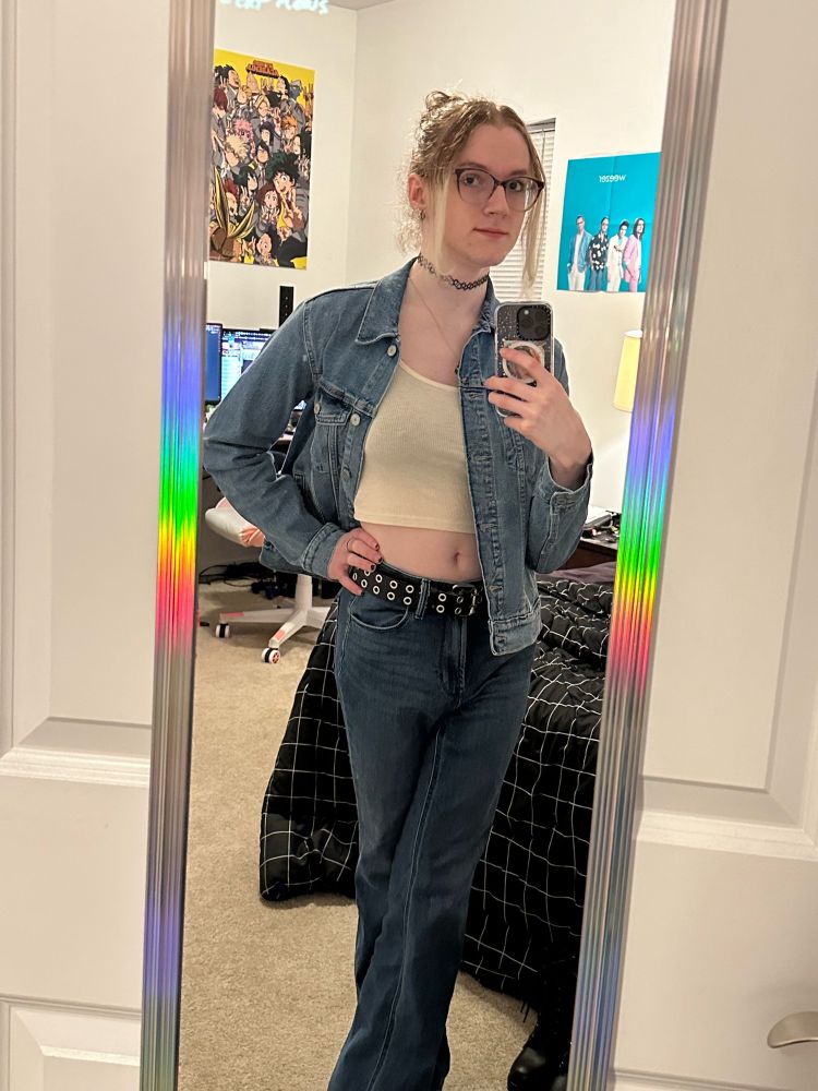 mirror selfie of kara in flared high waisted jeans, a cream crop top and a denim jacket. her hair is still straightened and up in a bun. she’s also wearing a tattoo choker, a black double grommet belt and gold necklace with a color by addition venn diagram on it as well as gold accented white flower earrings along with her normal mini black hoops. she has her legs cross and one hand on her hip, she seems a little disinterested but that’s just cause she’s still hungry and about to dig in to a breakfast bagel
