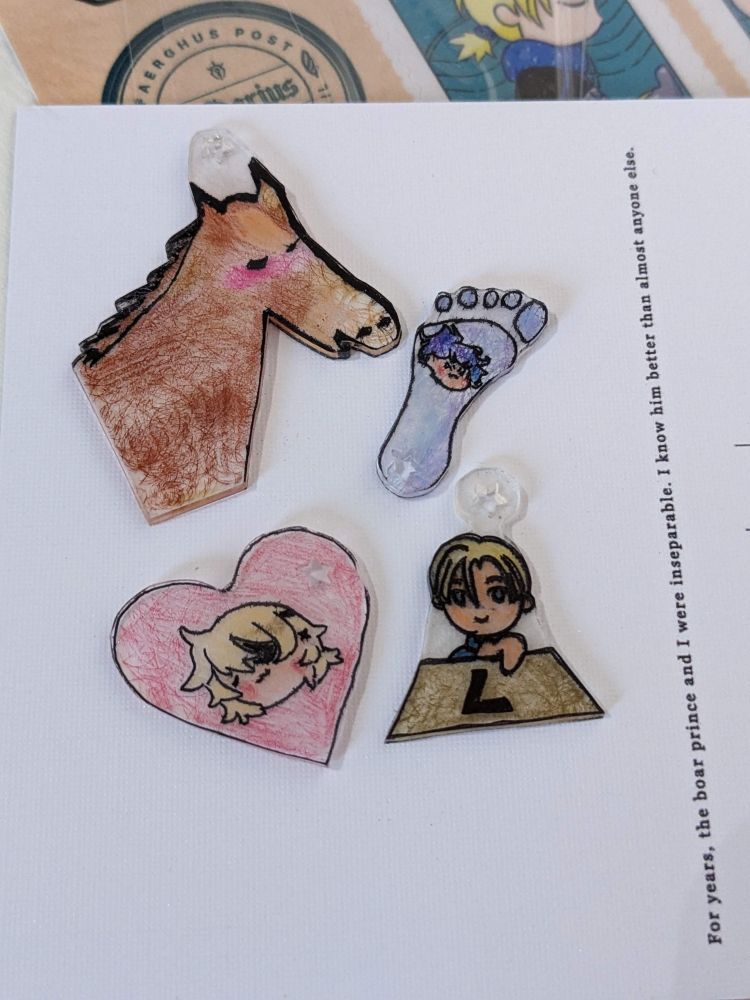 four shrinky dink charms: a blushing horse, a foot featuring Felix, a heart featuring Dimitri, and pre-skip Dimitri handing you an L