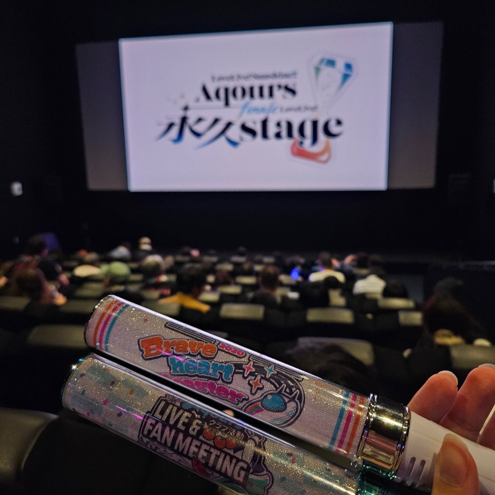 photo from a cinema screening of Aqours Finale Live at the Shinjuku Wald9, June 2025. two aqours concert penlights are held up in the foreground. 