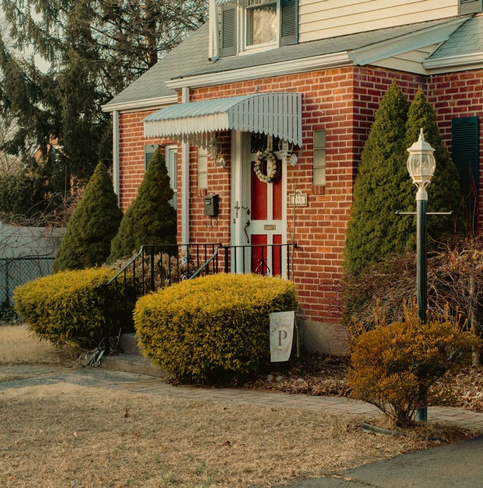 Image of a brick house with a red door, black hand rails and two lush green bushes on each side of the rails. A bit to the right is a black pole with an old style white lamp top.
