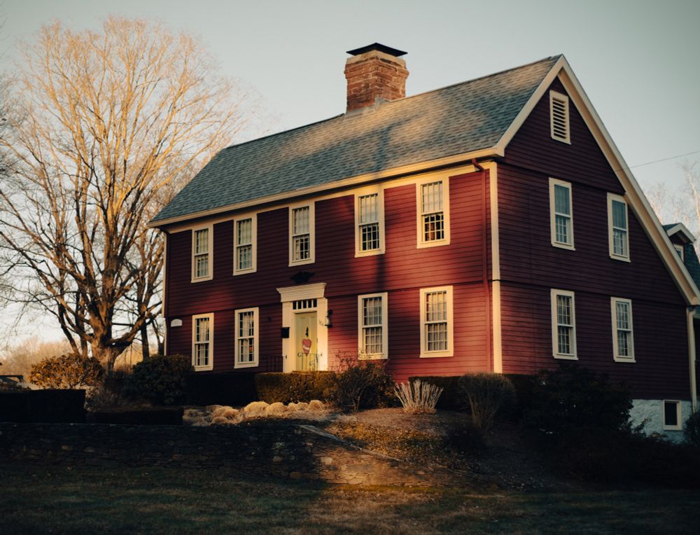 A classic New England farmhouse, like from the 1700s. It is maroon in color with white door and window frames. A brick chimney sits in the middle of the roof. On the front door is a heart that says "love".