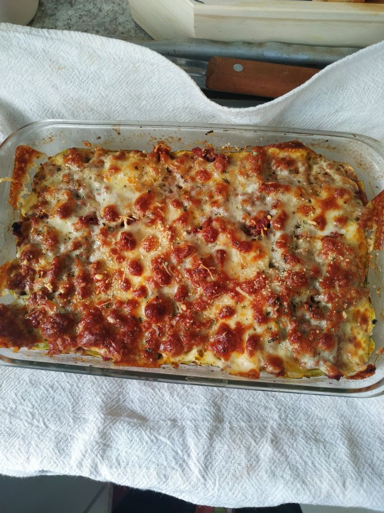 Imagem de uma travessa de vidro contendo uma lasanha de abobrinha, recheio de guisado e queijo gratinado por cima. Embaixo da travessa, um pano de prato branco