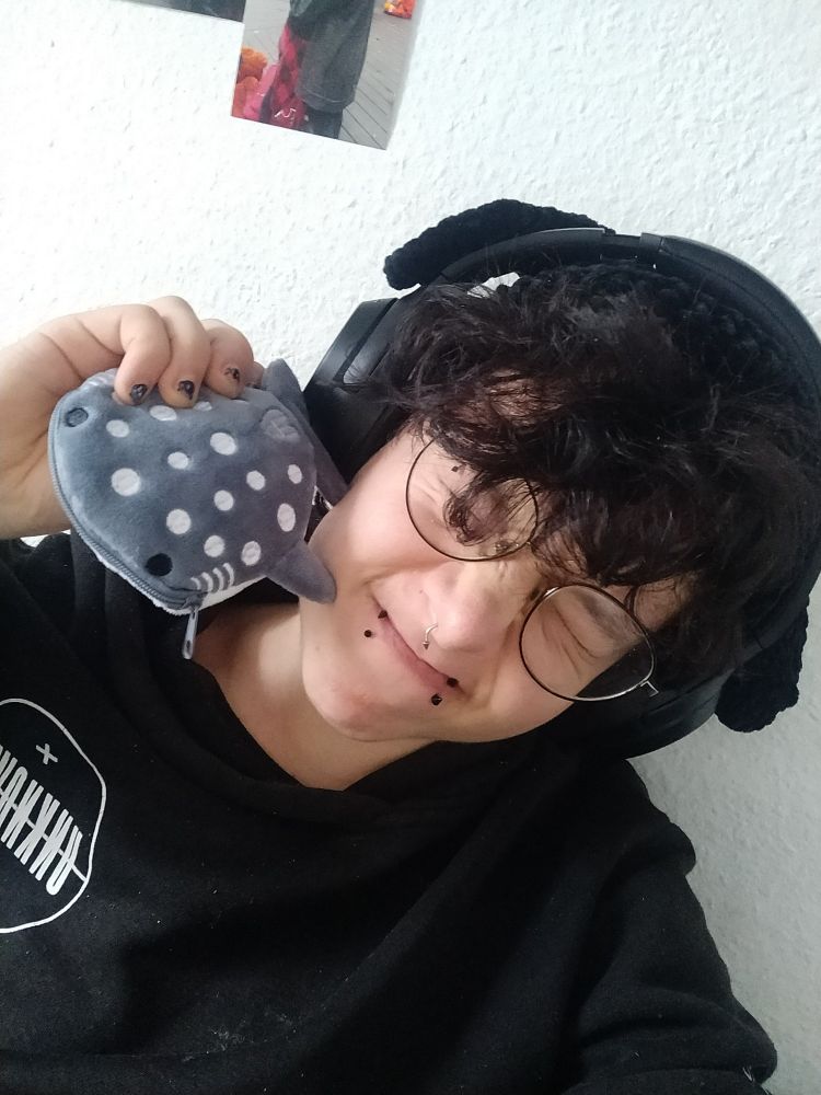 ein Selfie von mir (ich trage eine schwarze gehäkelte Hasenmütze, Kopfhörer und kneifen mein Gesicht zusammen), ich halte eine kleine Walhai Geldbörse neben mein Gesicht