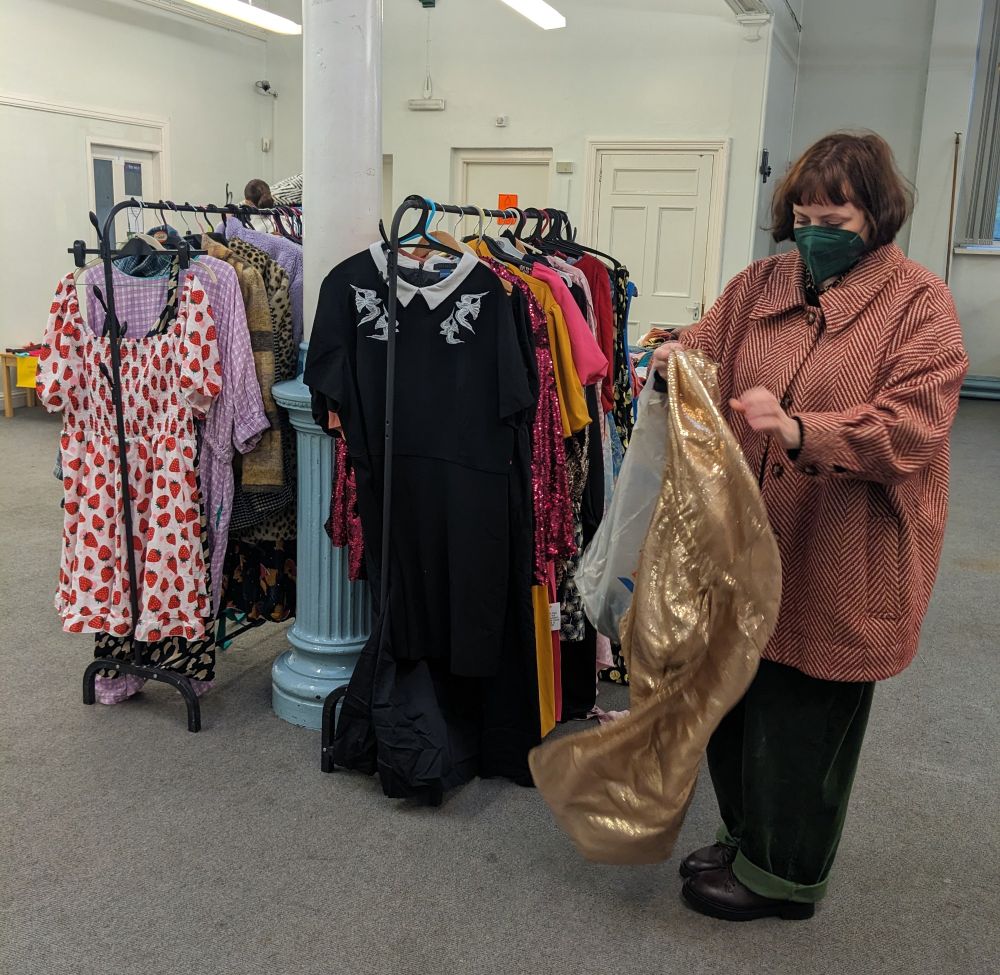 Two racks of clothes, showing many dresses including a white strawberry print milkmaid dress and a black dress with a white collar and lacework birds on it and my friend Jo, a white woman with brown hair, wearing a green respirator mask, a red tweed coat and green trousers, and holding a gold sequin dress 
