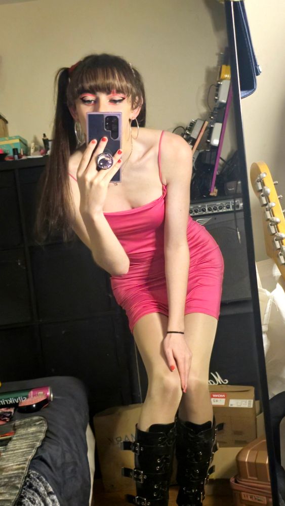 Shattered Polaroids taking a mirror selfie. Her phone is in front of most of her face but some neon pink eye makeup is still visible. She's wearing a very short pink dress, some shiny nude tights, and black boots. Her hair is tied in 2 ponytails and her fringe is flat across her forehead