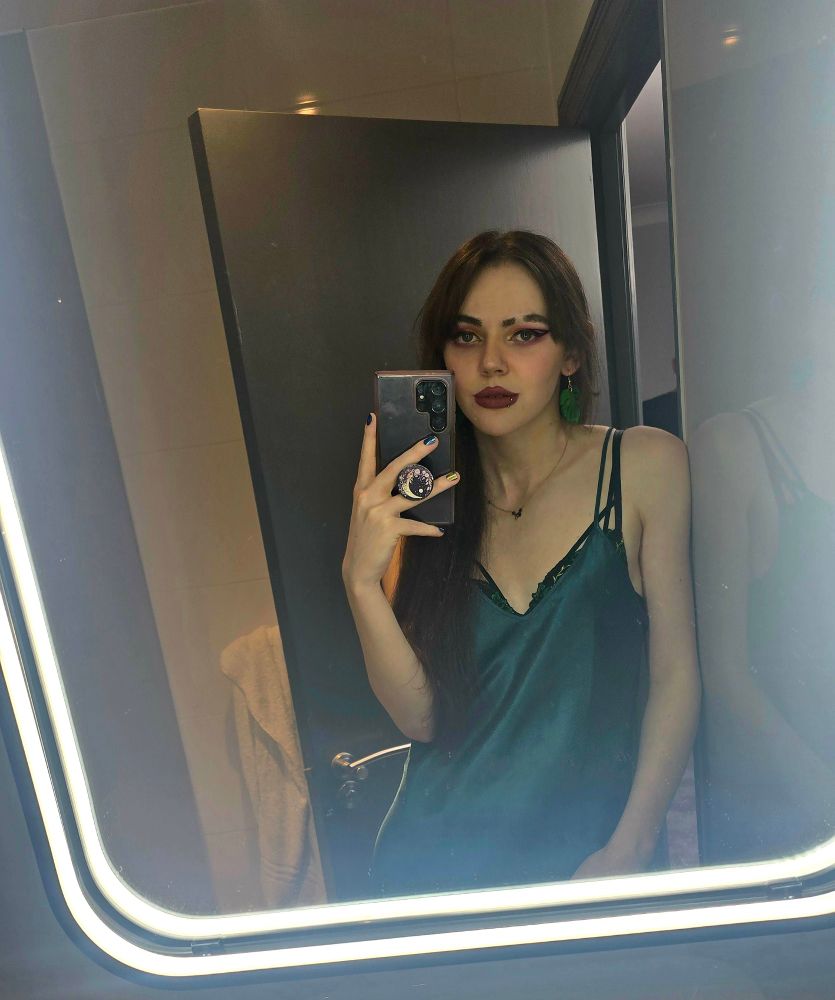 Shattered Polaroids taking a mirror selfie. Her long brown hair is down, she is wearing pink eyeshadow, her classic huge eyeliner wings, and a deep red lipstick. She's wearing a dark green satin slip. 