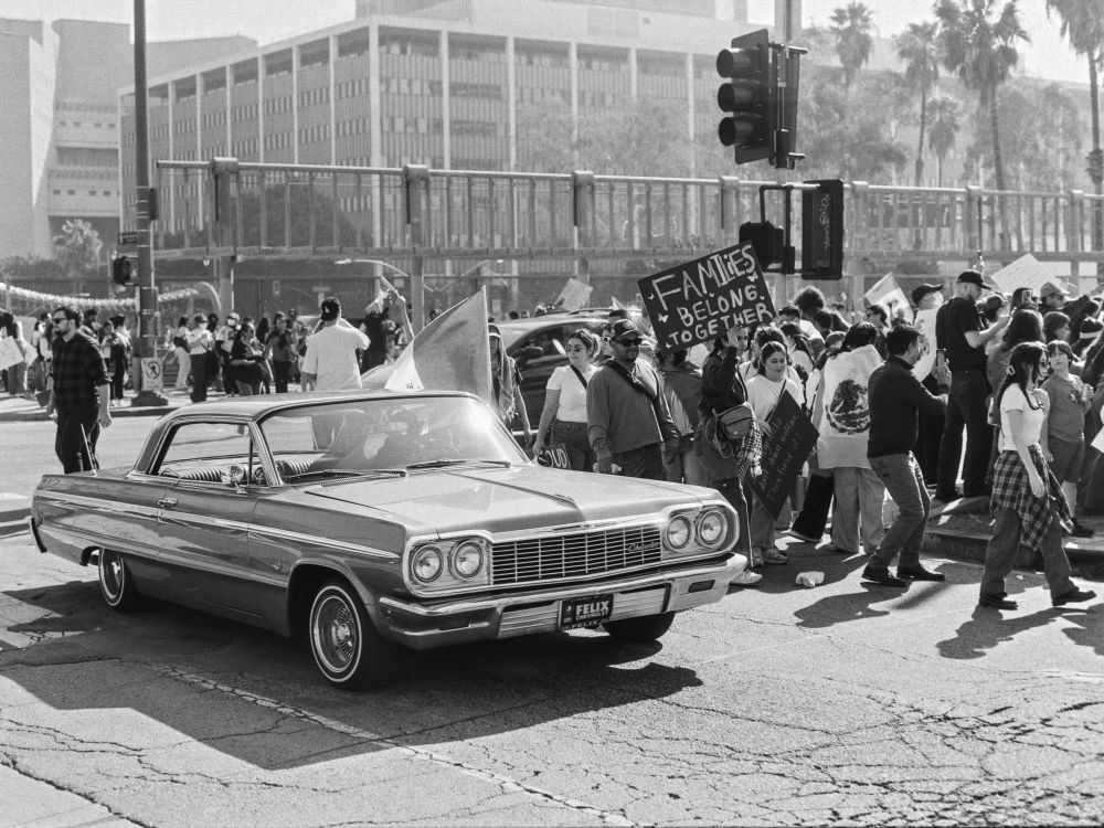 Protestors line the street holding anti-deportation signs while a classic low-rider car drives past. 