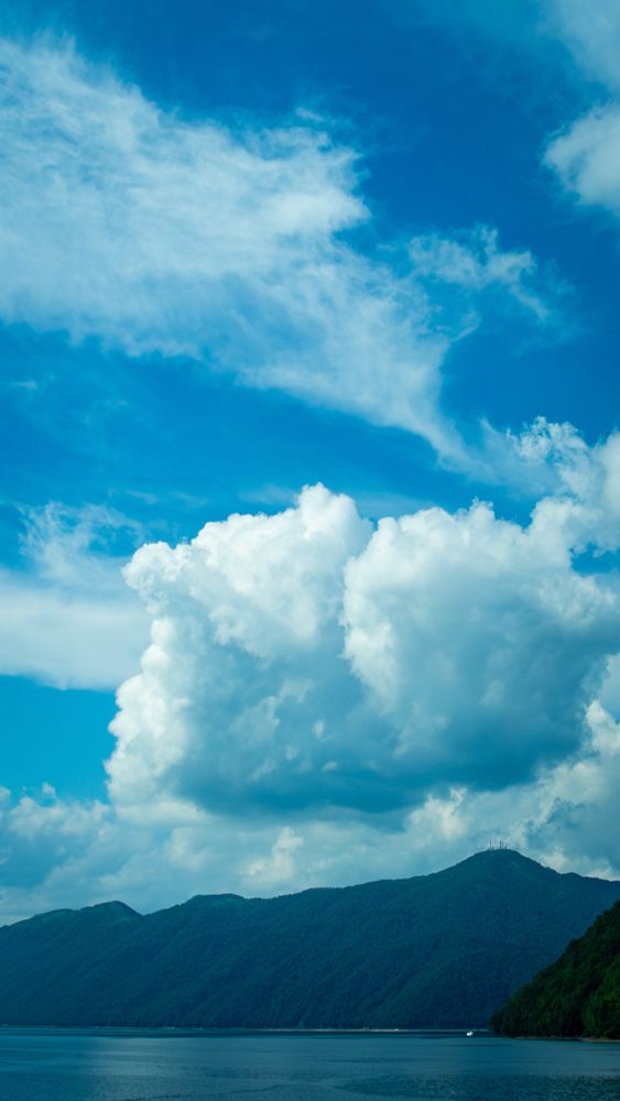 A scenic view of a calm lake bordered by forested mountains, with large, fluffy clouds and streaks of white against a deep blue sky. The colors and textures resemble those of an oil painting.
静かな湖と、それに沿って広がる森林に覆われた山々の風景。上空には大きな綿雲と白いすじ雲が浮かび、深い青空に映えている。色彩と質感が油絵のようにも見える。