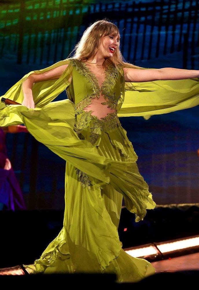 taylor swift on stage at the eras tour. she’s smiling and wearing a bright green dress. 