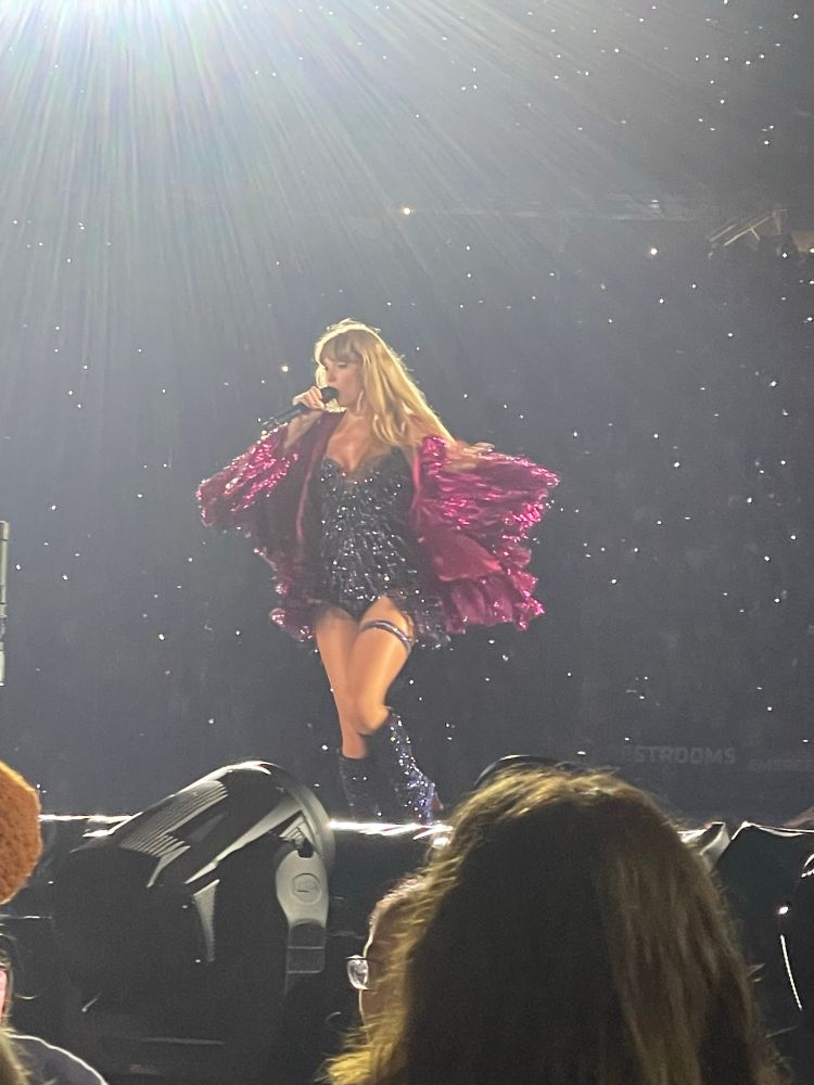 taylor swift performing karma at the eras tour. she’s wearing a blue sparkly bodysuit and a magenta jacket. 