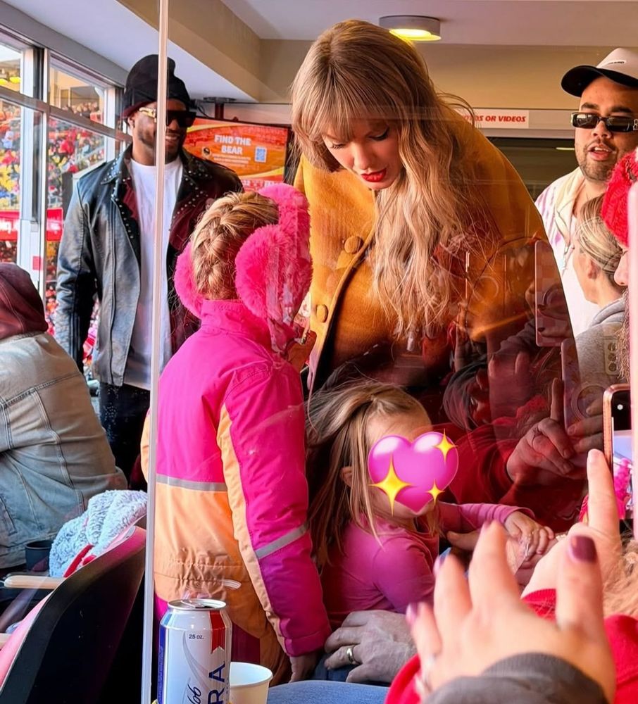 Taylor Swift wearing a fur coat and looking down. she’s in a suite and surrounded by people