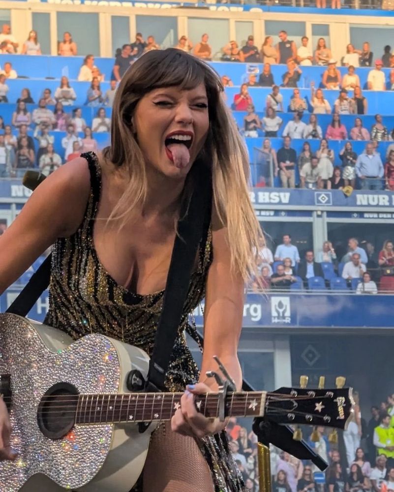 taylor swift performing at the eras tour, she’s playing guitar and sticking her tongue out