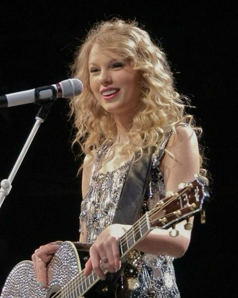 taylor swift on stage during the fearless tour. she’s wearing a silver tassel dress and holding a silver bedazzled guitar. 