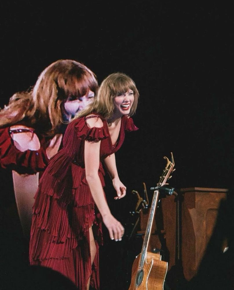 taylor swift on stage at the eras tour. she’s wearing a red dress with ruffles and reaching to pick up her acoustic guitar