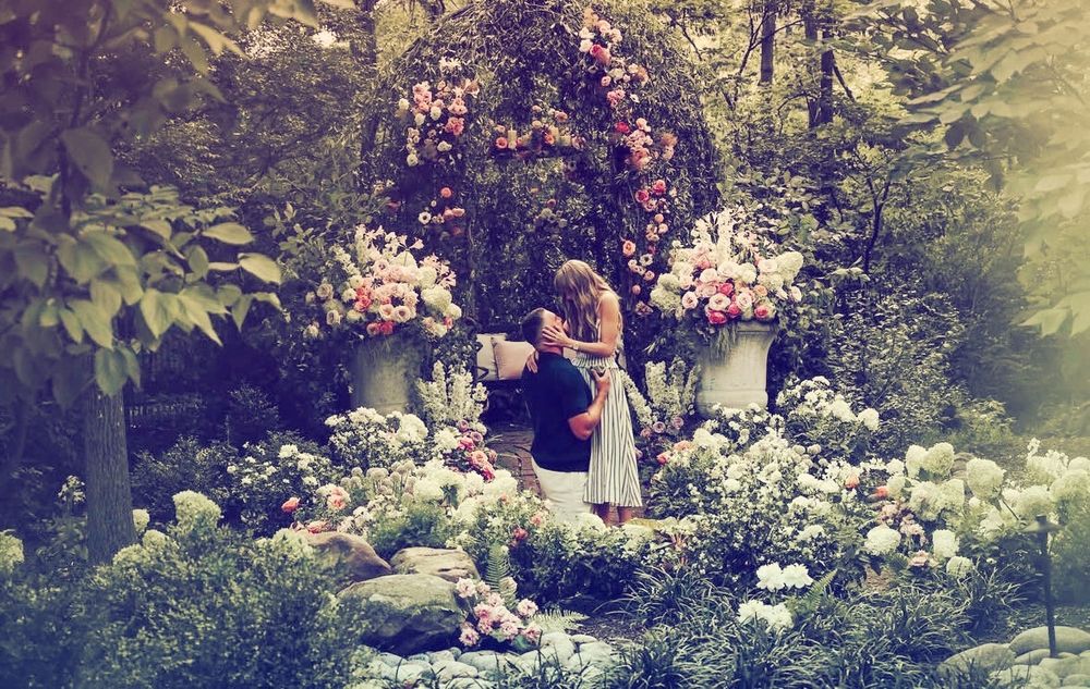 taylor swift and travis kelce in a garden. has on one knee, and taylor has her hands on his face. 