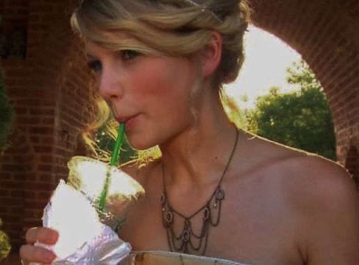 taylor swift with her hair in a bun wearing a strapless dress and dark dangling necklace. she’s sipping from a starbucks cup. 