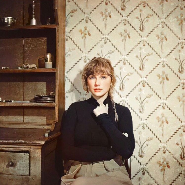 taylor swift photographed for her album evermore. she’s wearing a black turtleneck, and her hair is in a braid. she’s in front of beige floral wallpaper, and there’s a wooden dresser to her right. 