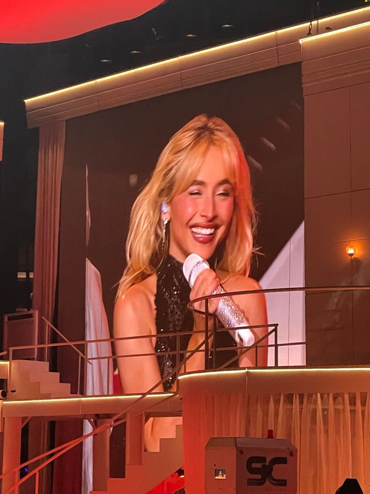 sabrina carpenter on the big screen, smiling at the crowd. she’s wearing a black bodysuit and holding a silver sparkly microphone. her stage set is partially obstructing the picture. 