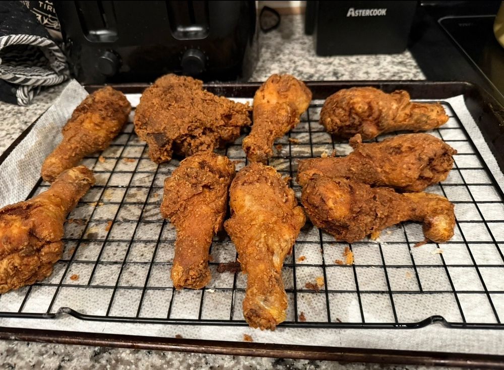 Picture of fried chicken on top of a baking rack, paper towel and cookie sheet. In the background a toaster is seen and a marble countertop. 