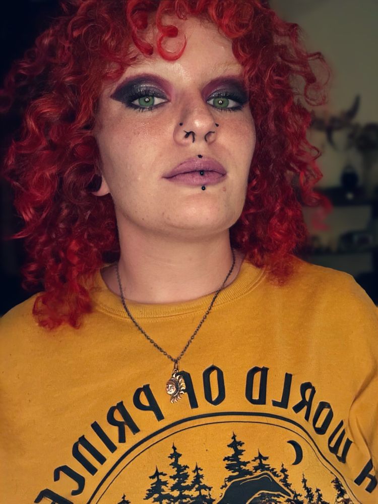 Close-up of the Asterin, showing her bleached brows, purple smokey eye, nose and lip piercings, and a sun pendant necklace. Shes in a bright orange sweater with black font and art displayed on it. 
