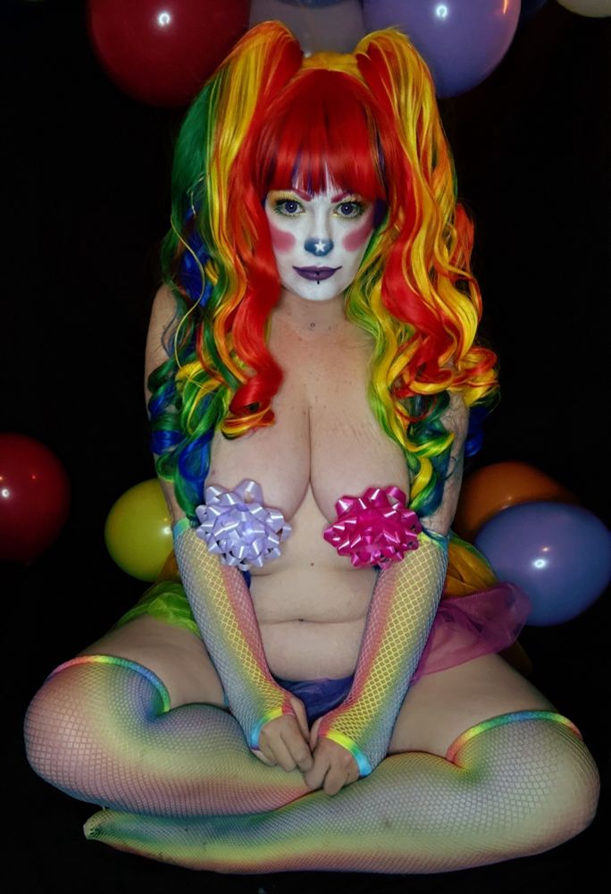 Goddess Asterin poses seated and topless, wearing only pastel bows over her chest. Her vibrant rainbow wig and makeup are paired with rainbow fishnet gloves and thigh-highs. She’s framed by balloons and layered tulle in pink, purple, and green hues.