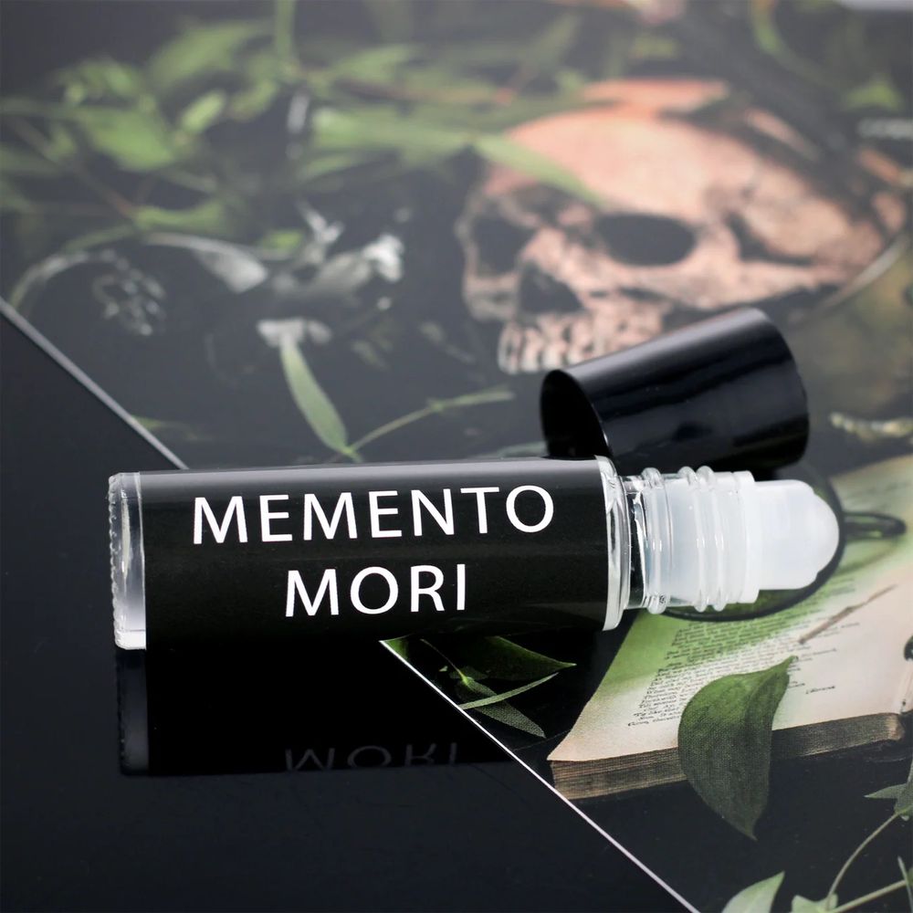 A small black perfume roller labeled “Memento Mori” in bold white text. The cap is off, revealing the clear rollerball tip. It rests on a dark background with greenery and a faint skull image. 