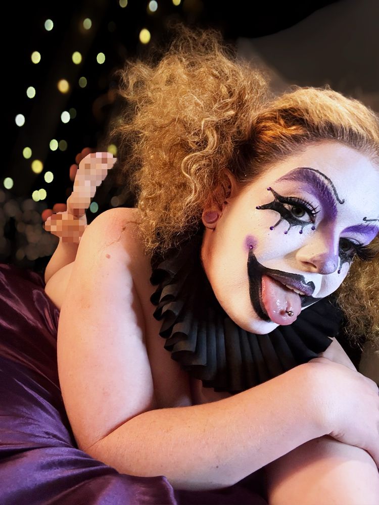 Asterin sticks out her tongue, while lying on her side, hair wild and face close to the camera. Her clown makeup is the same bold purple and black theme with piercing detail. Her tongue piercing is visible, and she wears the same black pleated collar.