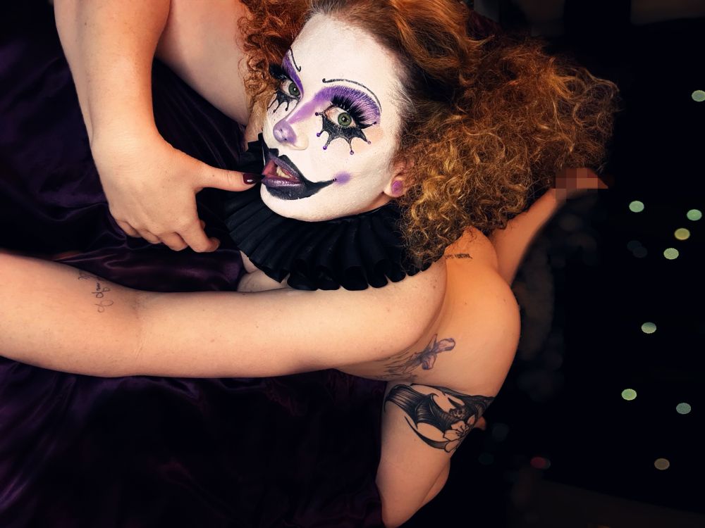 Goddess Asterin lies on purple satin sheets, fully nude with body parts obscured. Her face is painted in detailed clown makeup with bold purple and black eyeshadow, white base, and bold lashes. She wears a black collar and her curly red-blonde hair is styled in pigtails. She’s biting one nail and looking up.