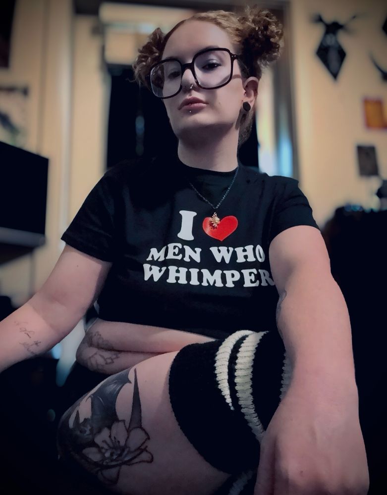 A dominating self-portrait of Goddess Asterin sitting in a dimly lit room. She has a black crop top with bold white text reading, “I ❤️ Men Who Whimper,” black-rimmed glasses, a sun necklace, barely visible black shorts and striped thigh-high socks. Her expression is assertive with a slight smirk. Her visible tattoos are on her arm (a cursive font) leg (bat with skull flower) and stomach (flower). The background features a softly lit indoor space with gothic-inspired decor.