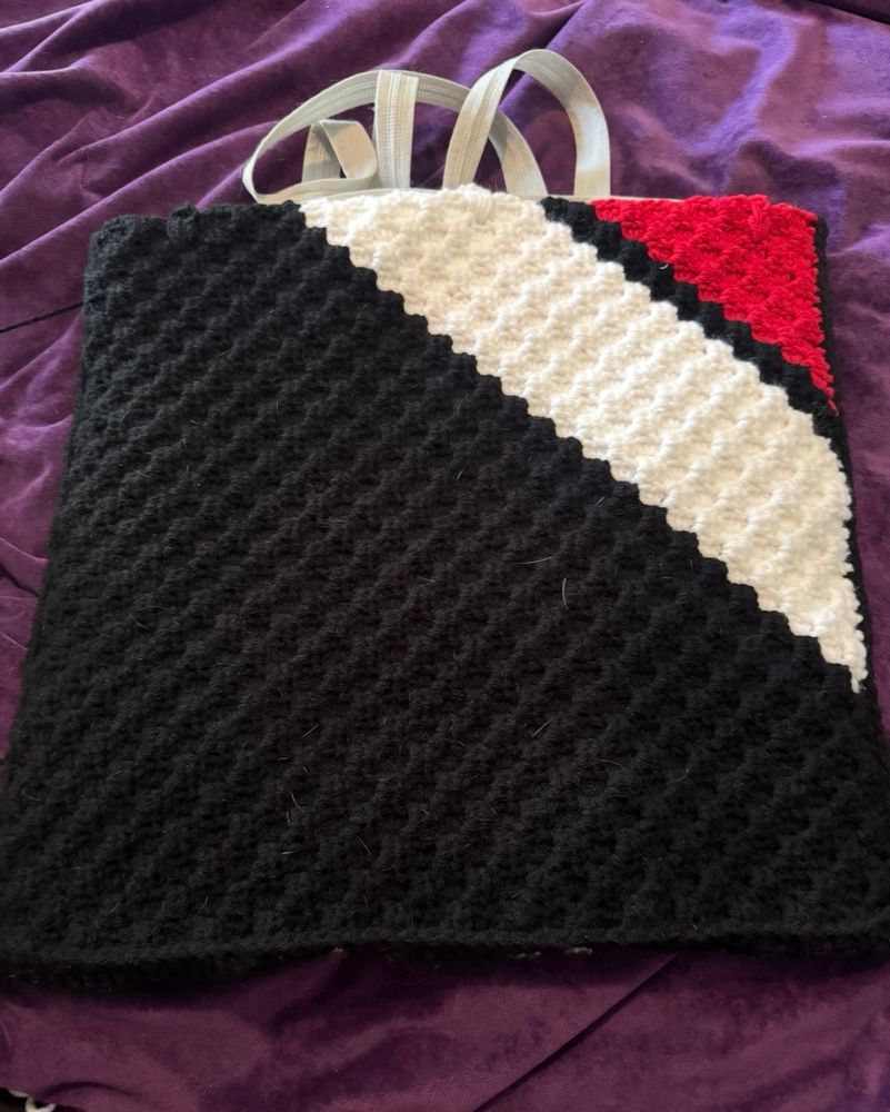 The reverse side of the crochet project, showing a textured corner-to-corner pattern divided diagonally into three color sections: black covering most of the square, white in the upper corner, and a small red triangle in the top right. Grey handles are attached at the top, and the piece lies flat on a purple cloth background.
