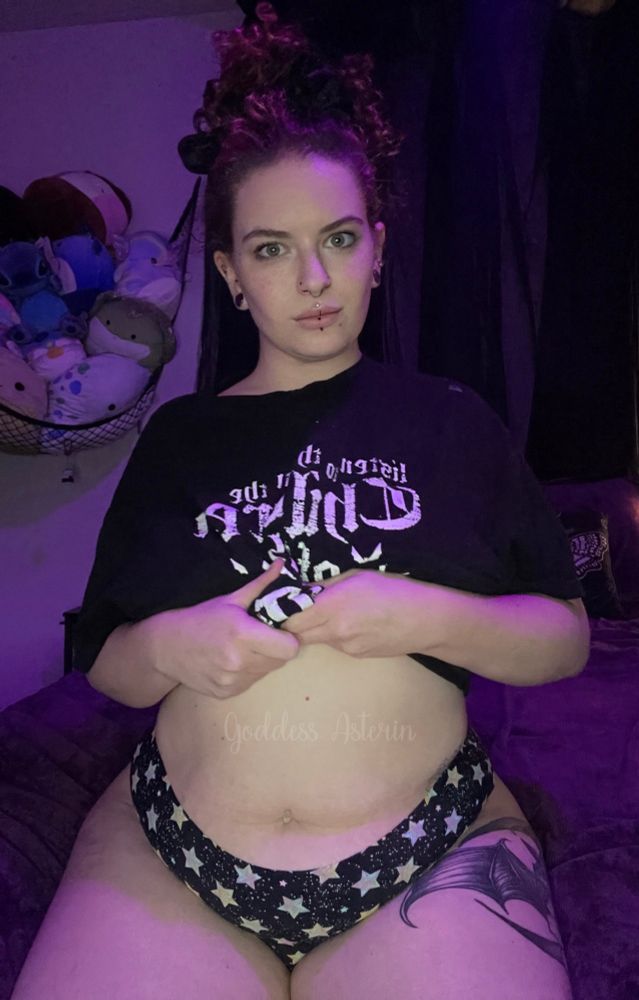 Goddess Asterin sits on a bed in a dimly lit room with soft purple lighting. She’s wearing a black graphic shirt and black underwear patterned with white and yellow stars. Her curly hair is tied up with a black scrunchie, and she has piercings on her lip and nose.