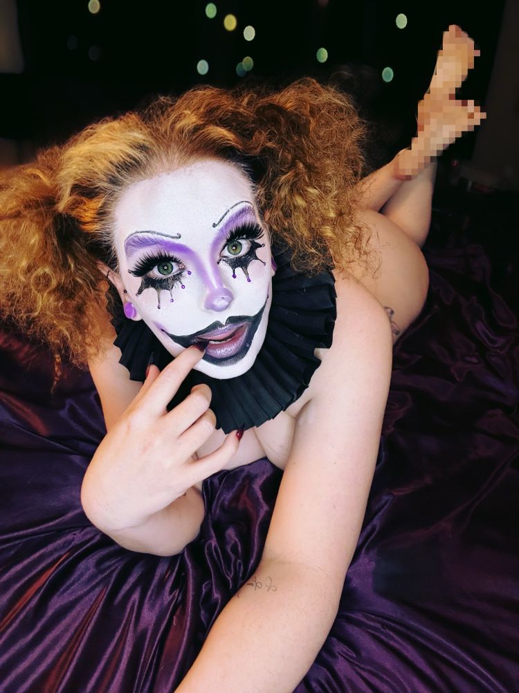 Asterin is lying on her stomach on purple satin bedding, propped up on her elbows with her feet in the air, face painted the same with clown makeup. One finger rests gently on her lips. Her curly hair in the same pigtail’s. 