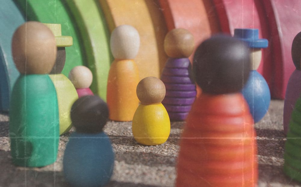 Un grupo de figuras de madera de la colección “Together” de Grapat se reúne en un entorno lleno de color. Las piezas representan personas diversas, con distintos colores, tamaños y formas. En el centro, una figura amarilla más pequeña destaca entre otras figuras más altas. Al fondo hay un arcoíris de madera.