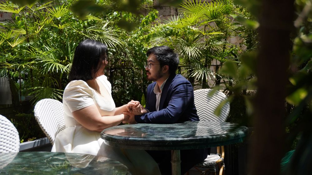 A couple sitting at a table in an outdoor garden, holding hands and looking lovingly at each other. They are framed with various greenery.