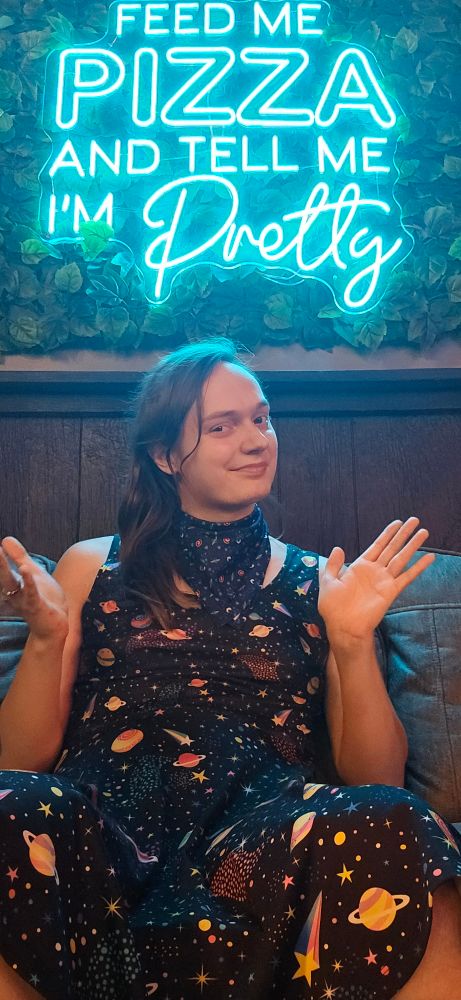 Neon sign on leaf background that says "feed me pizza and tell me I'm pretty"
Below: Sparkle, a trans woman with brown hair, brown eyes, wearing a space bandana and space themed dress sitting on a couch pointing at the neon sign