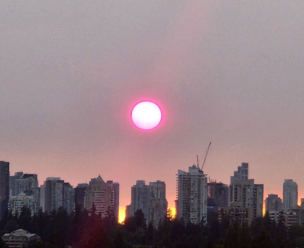A red sun in a hazy sky hangs over a row of apartment towers at dusk.