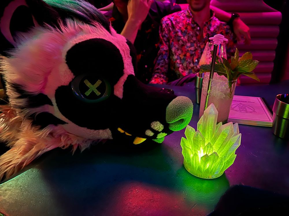 Hyena fursuit headpiece on a table next to a glowing green crystal cluster and a cocktail glass