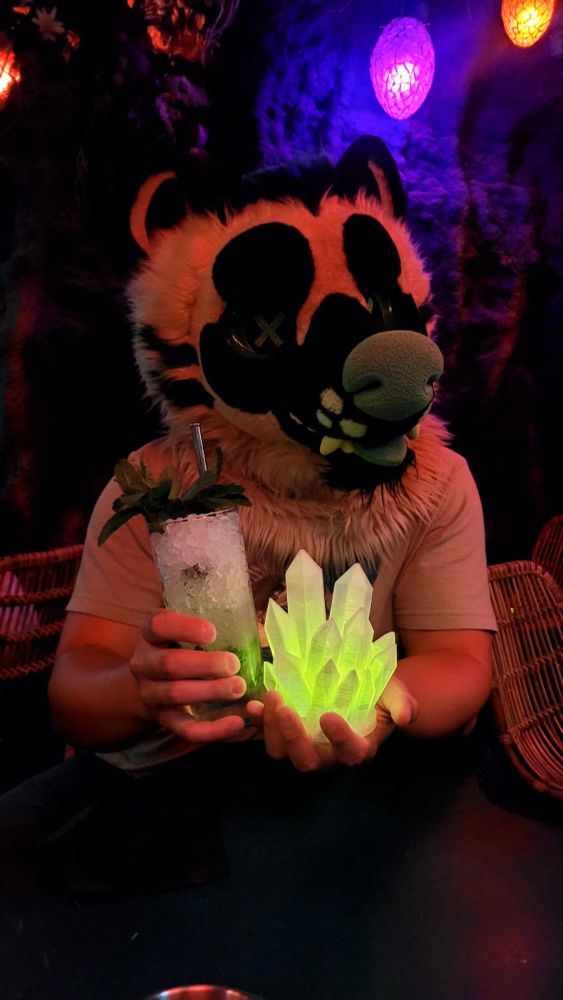 Hyena fursuiter holding a green glowing crystal cluster and a tall cocktail full of crushed ice and garnished with mint