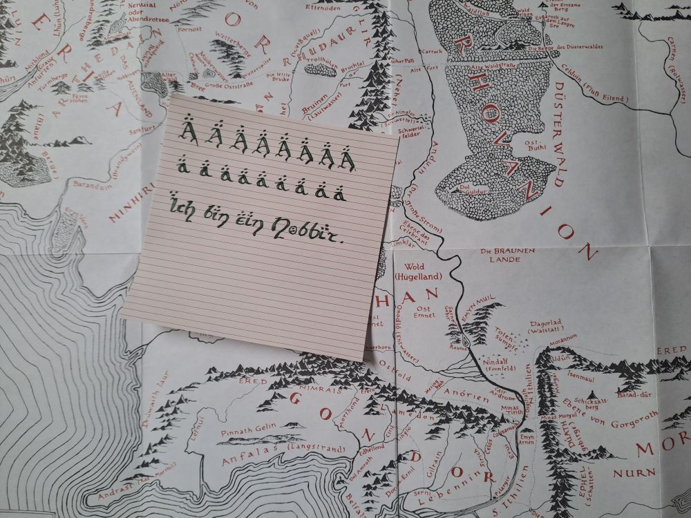 Je eine Zeile „A“ und „a“. Darunter steht noch: Ich bin ein Hobbit. Alles wurde in grüner Tinte in Hobbit-Schrift geschrieben. Der Zettel liegt auf einer Mittelerde-Landkarte.