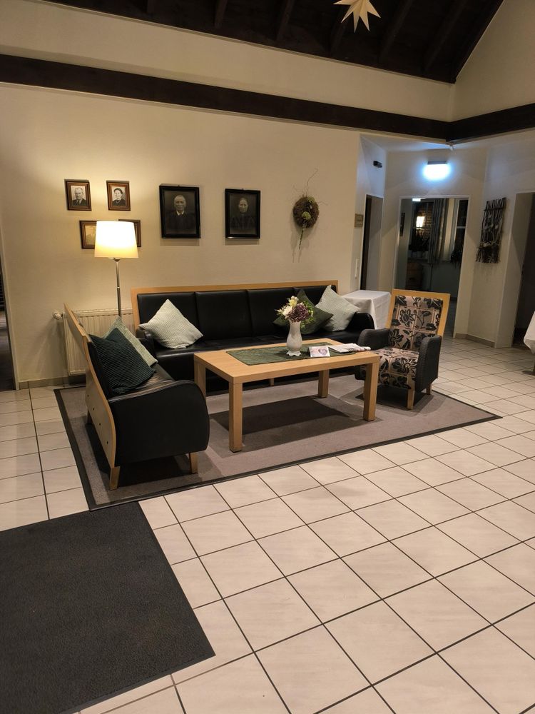 Gemütliche Sitzgruppe in einem hellen Eingangs- oder Aufenthaltsbereich mit schwarzem Sofa, zwei Sesseln, Holztisch und Dekoration wie Kissen, Blumenstrauß und Wandbildern älterer Personen.